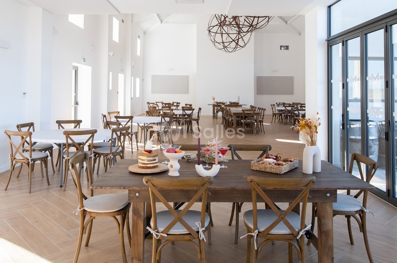 Salle lumineuse avec tables en bois, chaises croisées et décorations minimalistes, vue sur de grandes baies vitrées.
