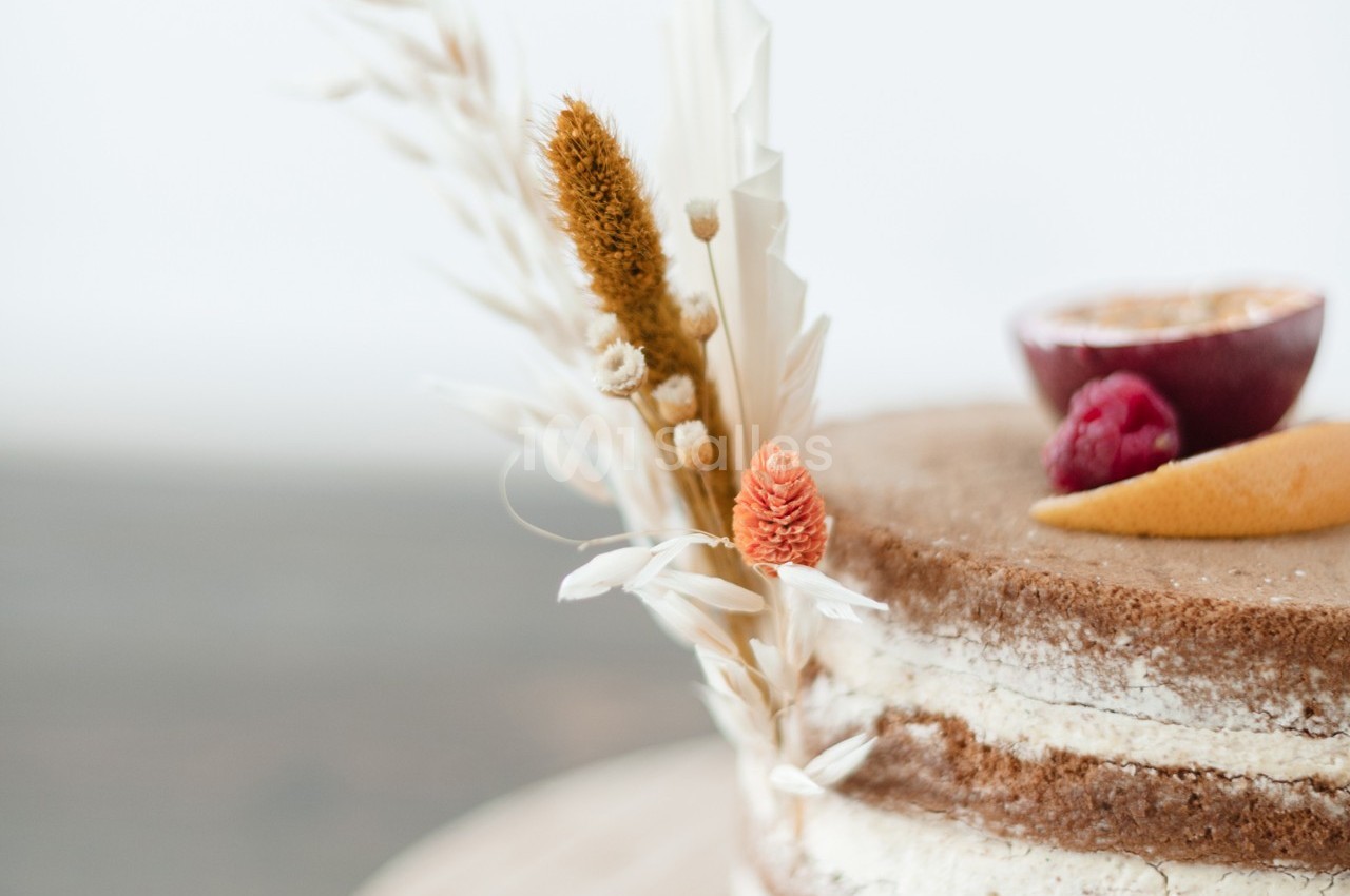Gâteau nu décoré de fleurs séchées, d'une tranche de fruit de la passion, d'une framboise et d'un zeste d'agrume.