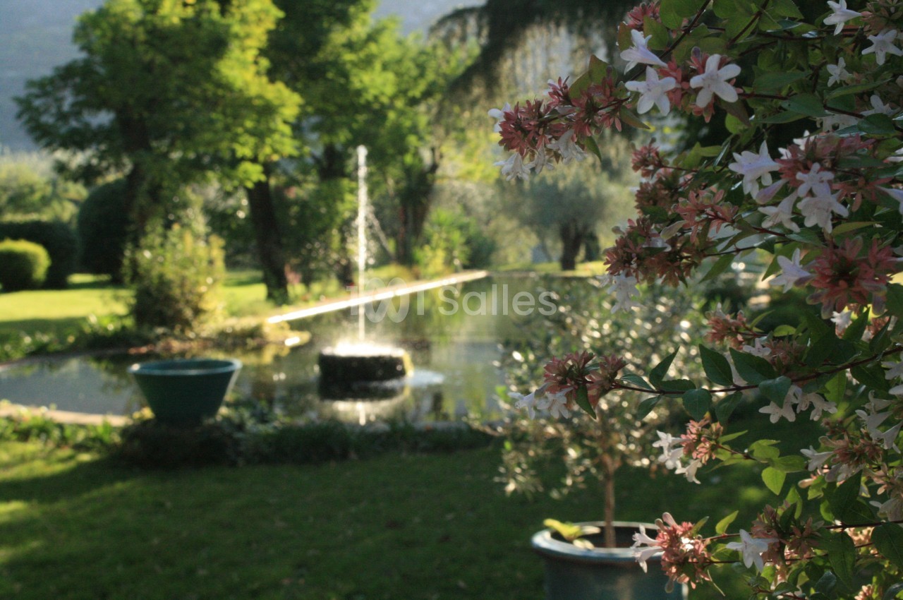 Vue d'un jardin avec un bassin, une fontaine centrale et des fleurs roses au premier plan.