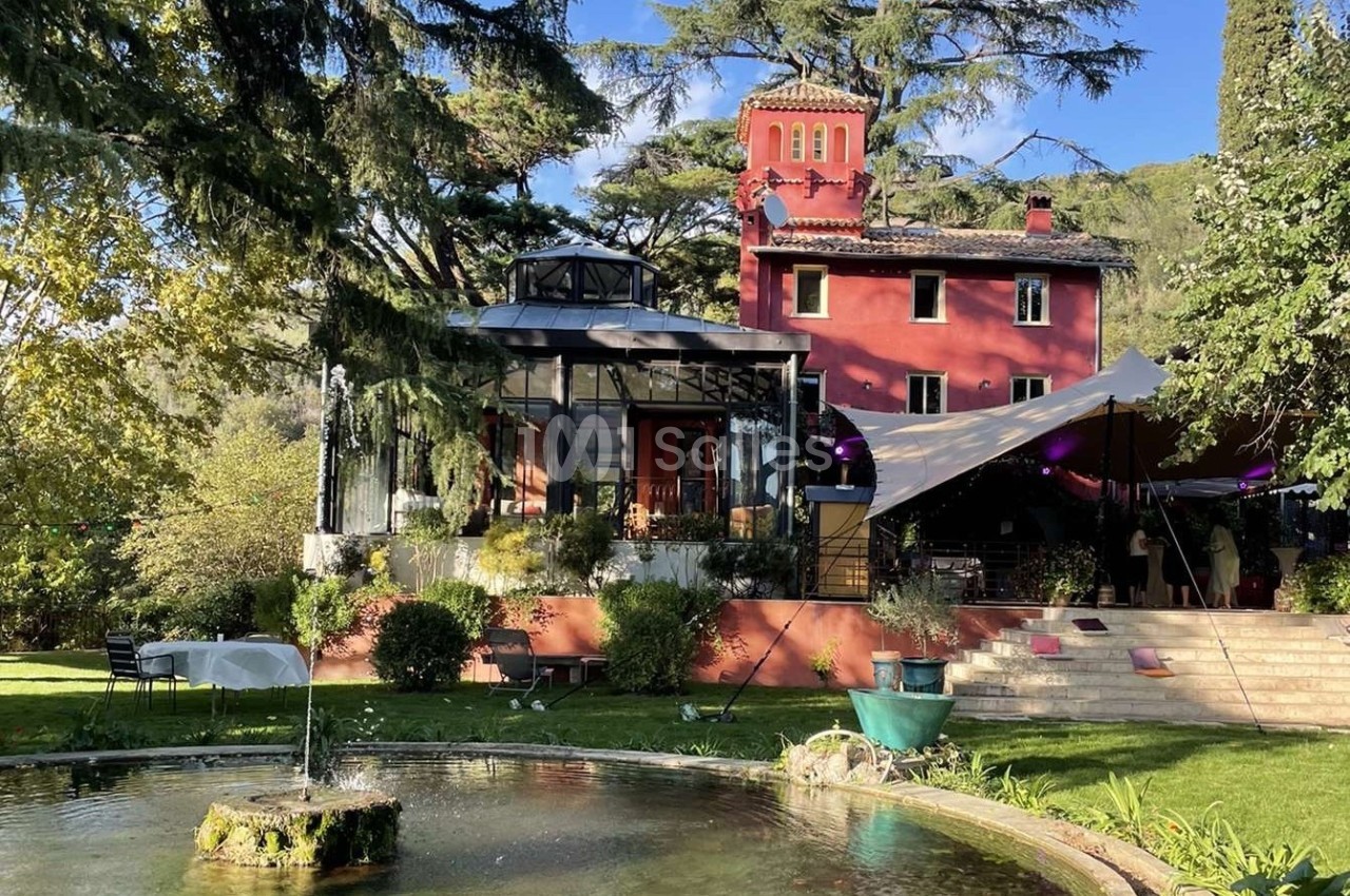 Grande maison rouge entourée de verdure, avec une verrière, une tente et un bassin au premier plan.