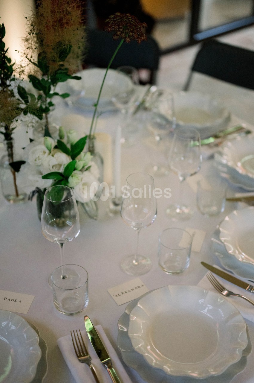 Table dressée avec vaisselle blanche, verres, couverts, bougie et bouquet de fleurs blanches et vertes.