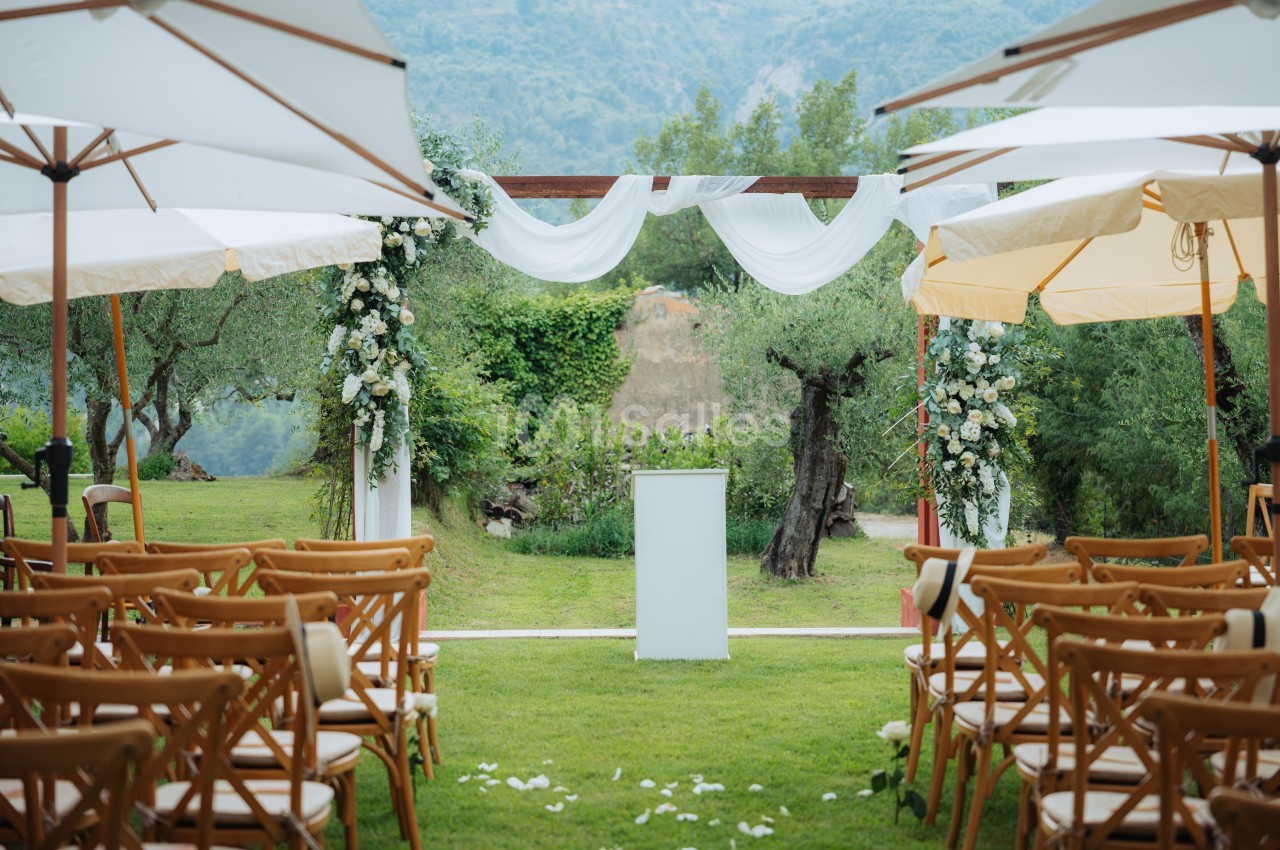Allée centrale décorée pour une cérémonie en plein air, avec chaises en bois, fleurs blanches et vue sur un paysage…