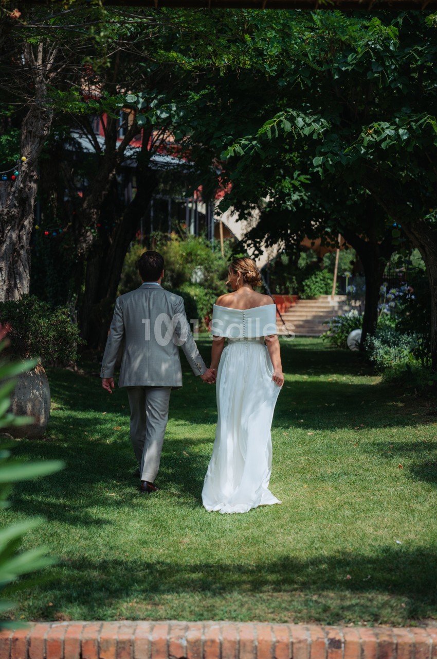 Un couple en tenue de mariage marche main dans la main dans un jardin verdoyant.
