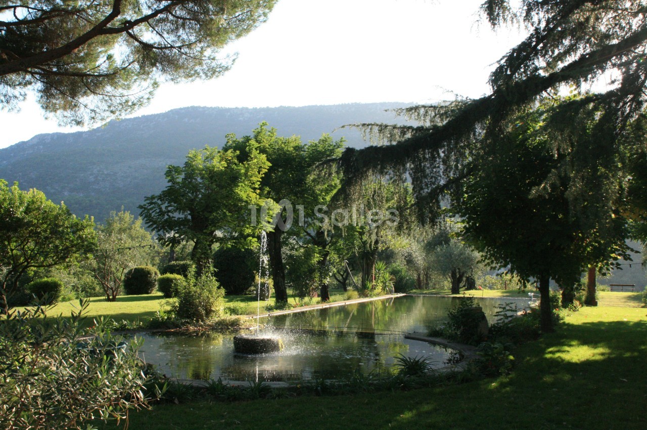 Étang circulaire avec une fontaine entouré de végétation luxuriante, sous un ciel clair et une montagne en arrière-plan.