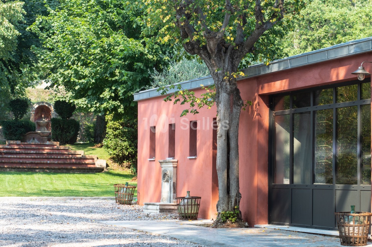 Bâtiment aux murs rouges avec grandes fenêtres, entouré d'arbres et d'une fontaine dans un jardin verdoyant.