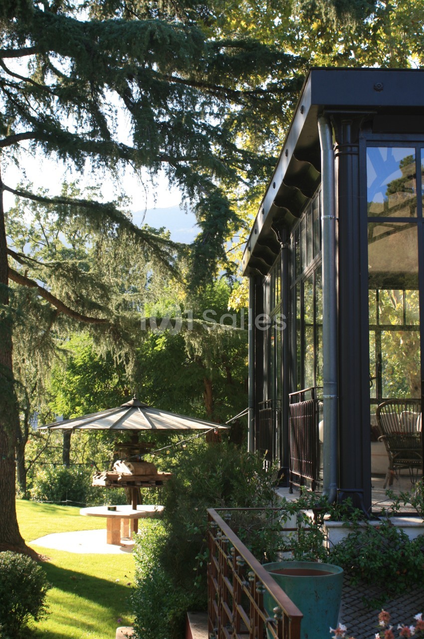 Véranda en verre et métal donnant sur un jardin arboré avec une table et un parasol sous les arbres.