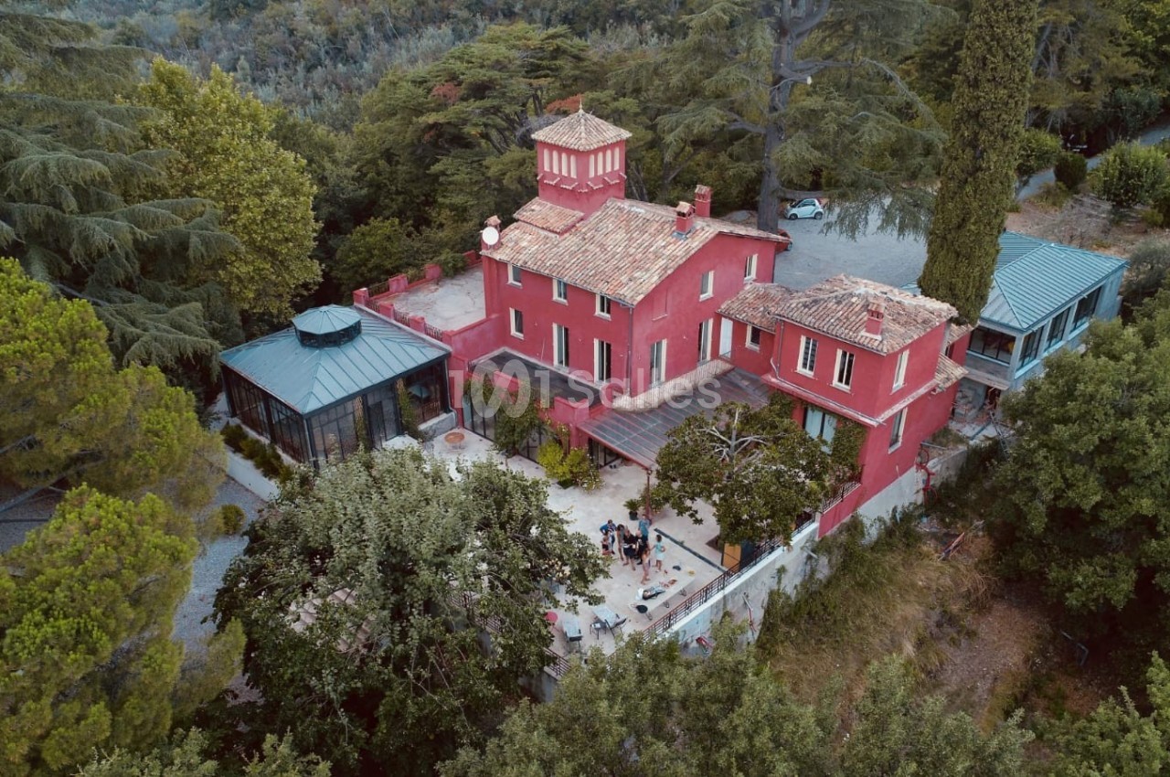 Vue aérienne d'une grande maison rouge entourée d'arbres, avec une terrasse et une verrière attenante.