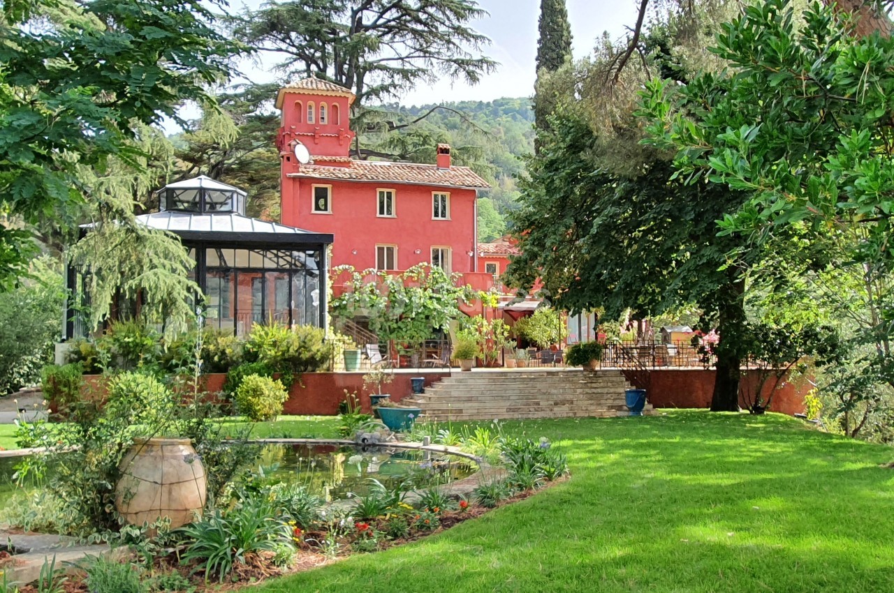 Grande maison rouge entourée de verdure, avec un jardin paysager, un bassin et des arbres matures.