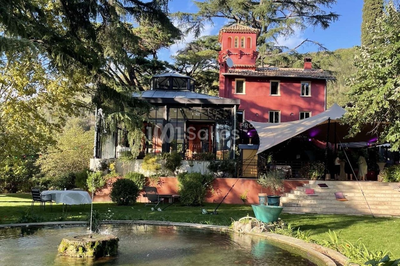 Grande maison rouge avec verrière, entourée d'arbres et d'un jardin avec une fontaine au premier plan.