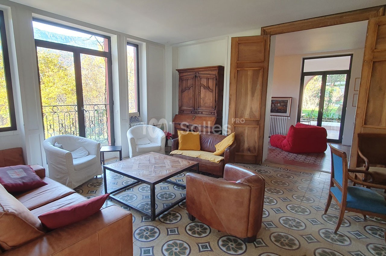 Salon lumineux avec canapés, fauteuils en cuir, table basse en bois, sol en carreaux décoratifs et grandes fenêtres.