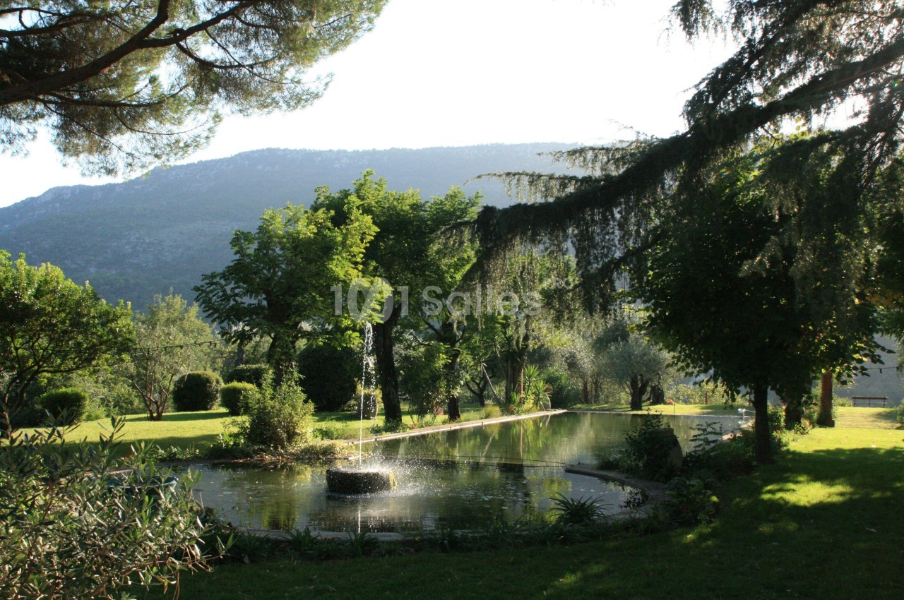 Fontaine entourée de végétation dans un parc verdoyant avec des arbres et une montagne en arrière-plan.