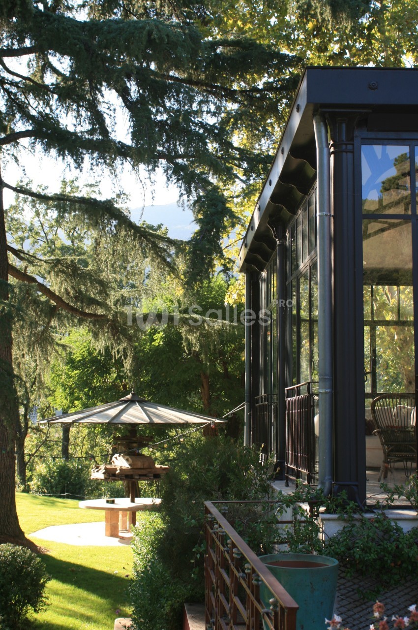 Véranda vitrée donnant sur un jardin verdoyant avec parasol et arbres en arrière-plan.