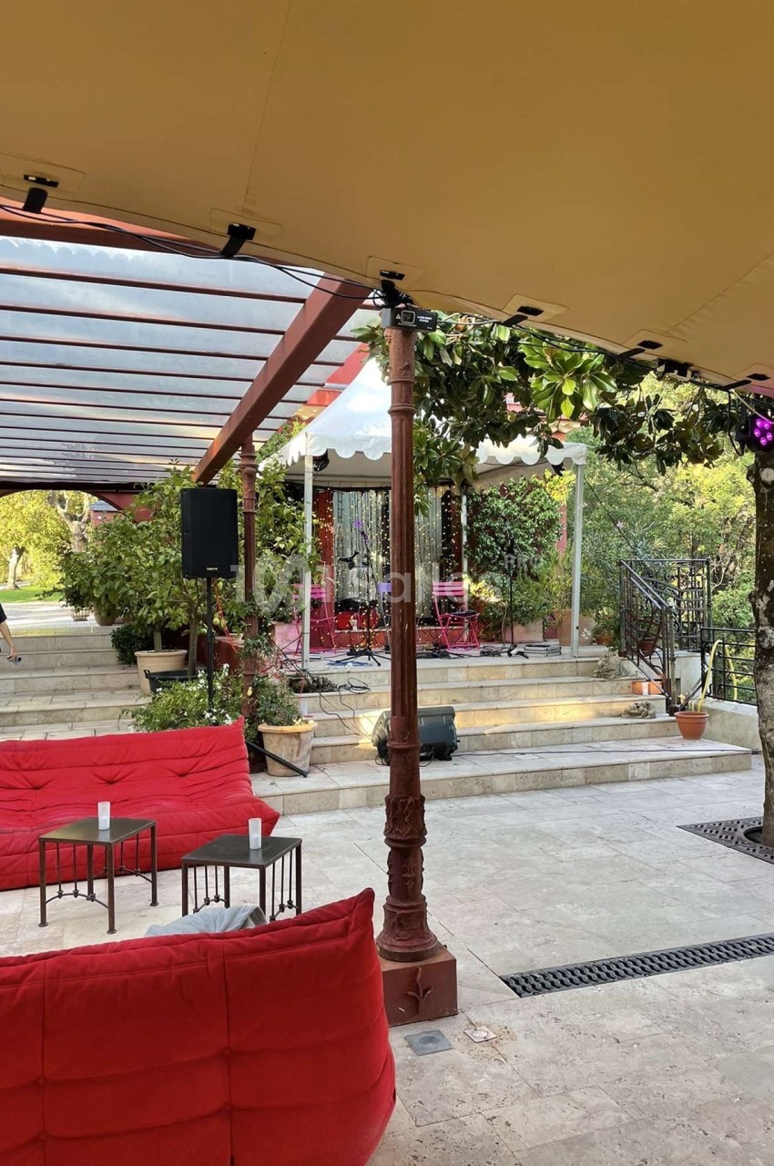 Terrasse extérieure avec canapés rouges, tables basses, scène équipée et végétation environnante sous une pergola.