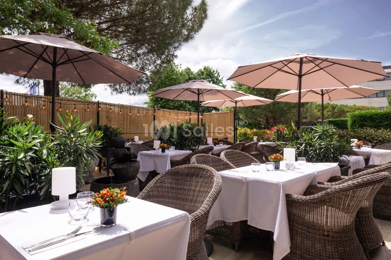 Terrasse d'un restaurant avec tables dressées, chaises en rotin, parasols et végétation environnante.