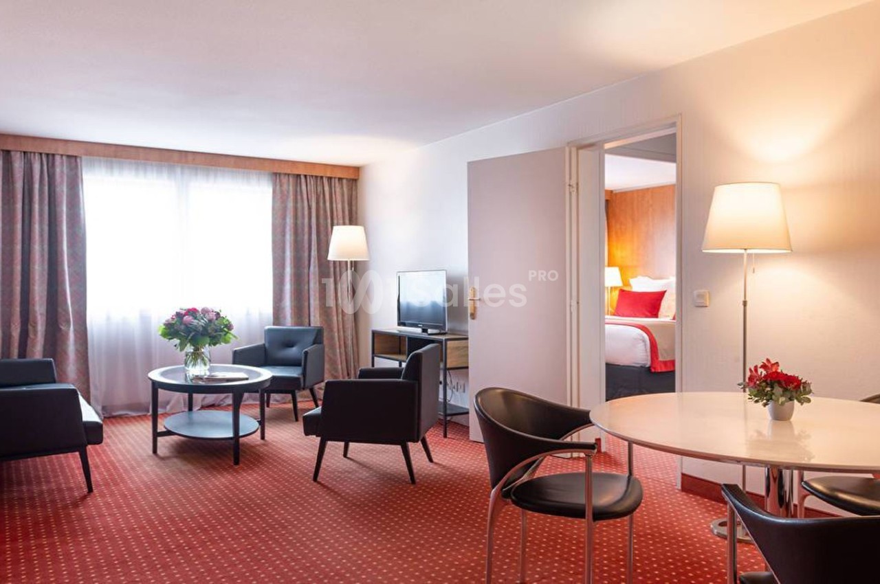 Salon d'hôtel avec fauteuils, table basse, table à manger, lampes et vue sur une chambre adjacente.