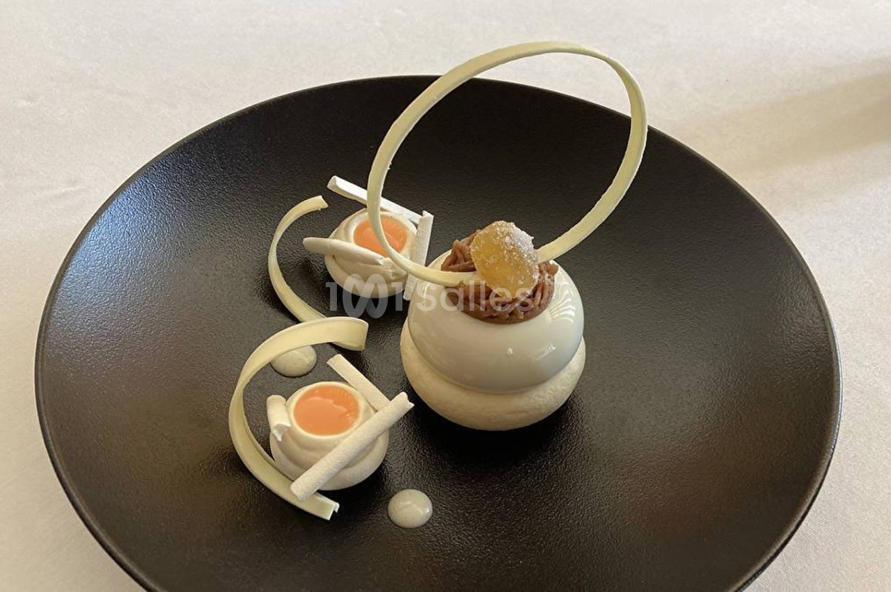 Dessert raffiné présenté sur une assiette noire, composé de mousse, décorations en chocolat blanc et garnitures élégantes.