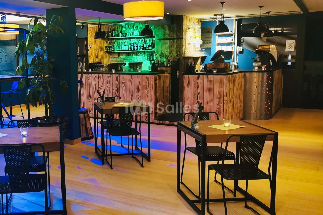 Salle de restaurant moderne avec tables en bois, chaises noires, éclairage chaleureux et un bar en arrière-plan.
