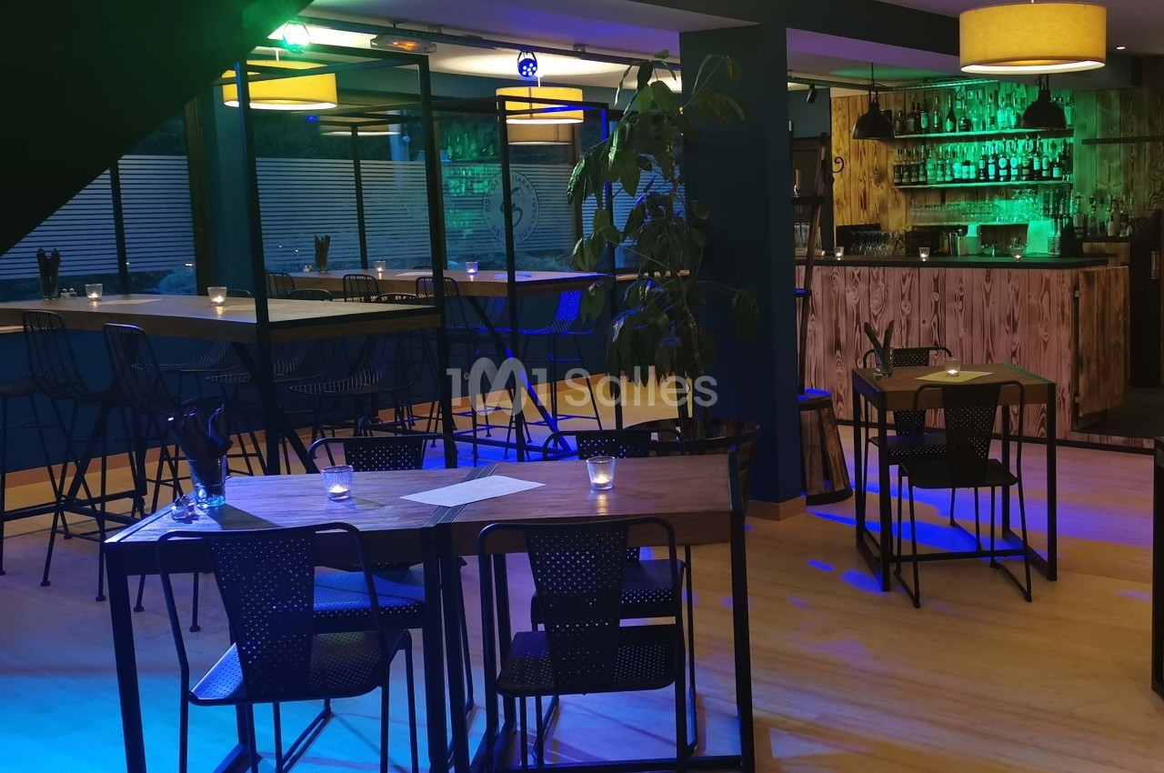 Salle de restaurant moderne avec tables en bois, chaises noires, éclairage tamisé et bar en arrière-plan.