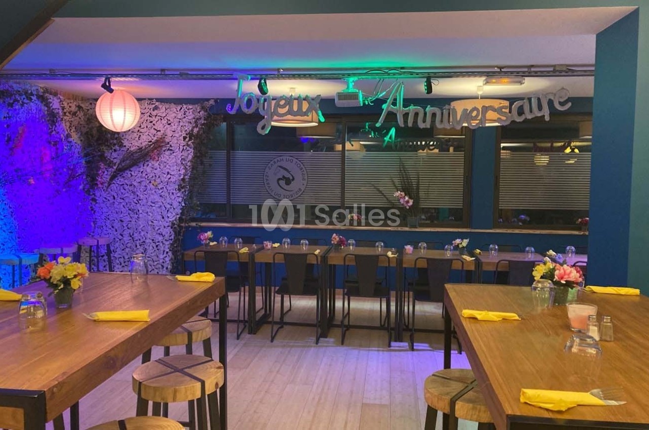 Salle décorée pour un anniversaire avec tables en bois, chaises hautes, fleurs et éclairage coloré.