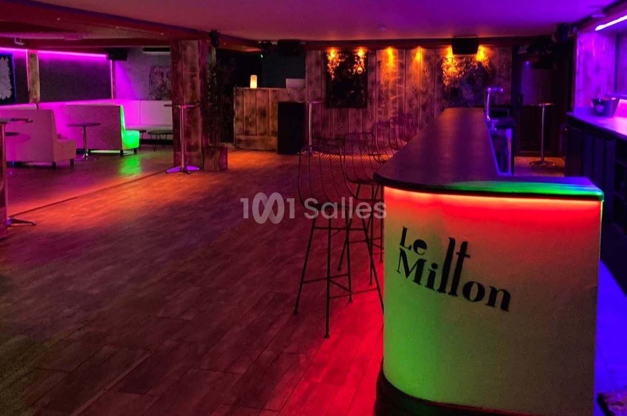Intérieur d'une salle avec bar éclairée par des lumières colorées, ambiance tamisée et espace aménagé pour des événements.