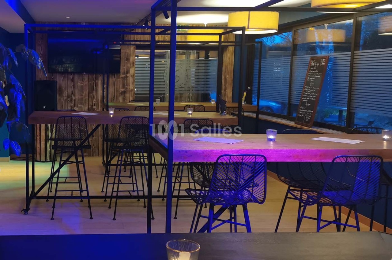 Salle moderne avec tables hautes en bois, chaises en métal, éclairage chaleureux et grandes fenêtres avec stores.