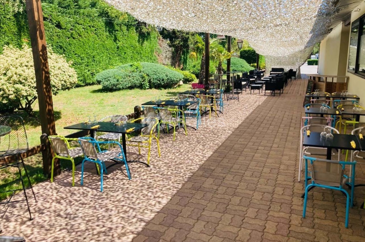 Terrasse extérieure avec tables et chaises colorées, ombragée par un filet, entourée de verdure.