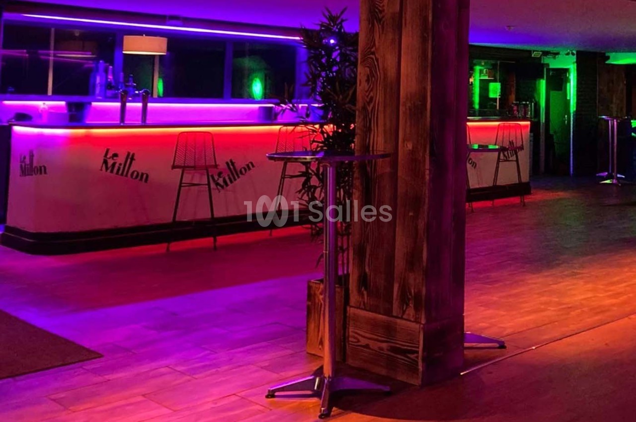 Salle de bar avec comptoirs éclairés par des lumières néon violettes et vertes, sol en bois et tables hautes.