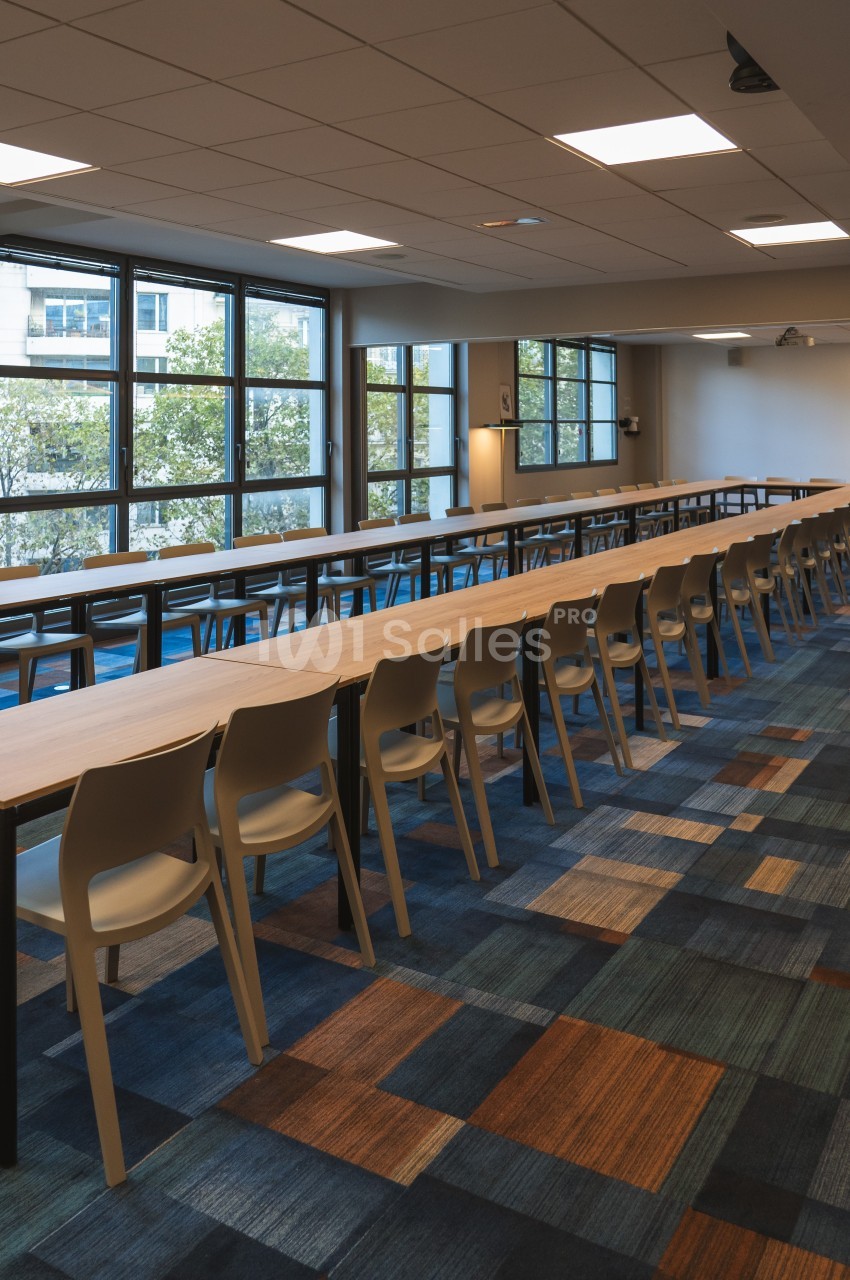 Salle de réunion lumineuse avec de longues tables en bois, des chaises alignées et un sol à motifs colorés.