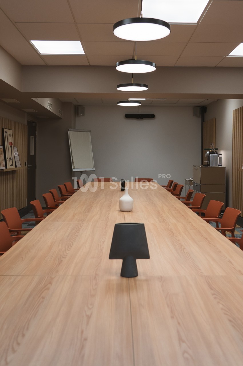 Salle de réunion avec une grande table en bois, des chaises orange, un tableau blanc et des cadres accrochés aux murs.