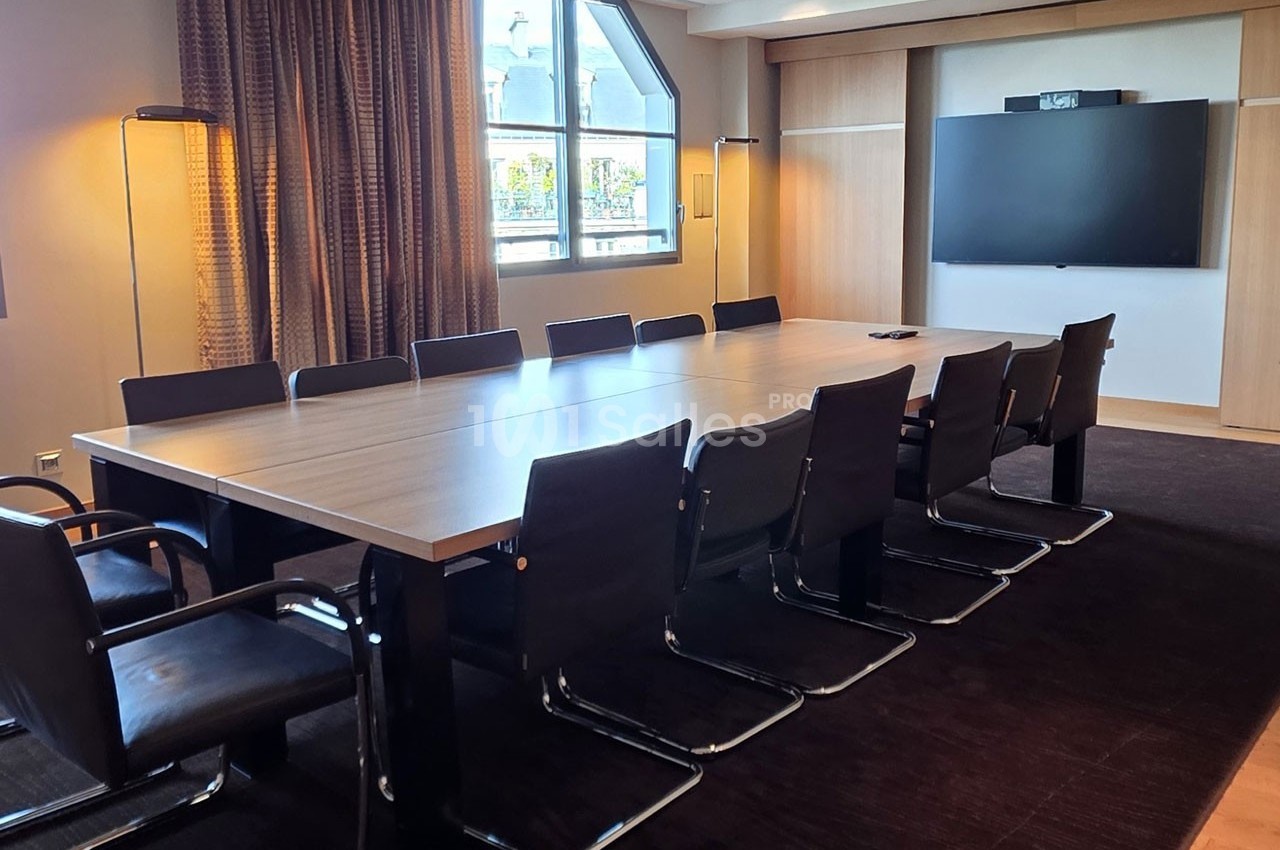 Salle de réunion moderne avec une grande table rectangulaire, des chaises noires et un écran mural.
