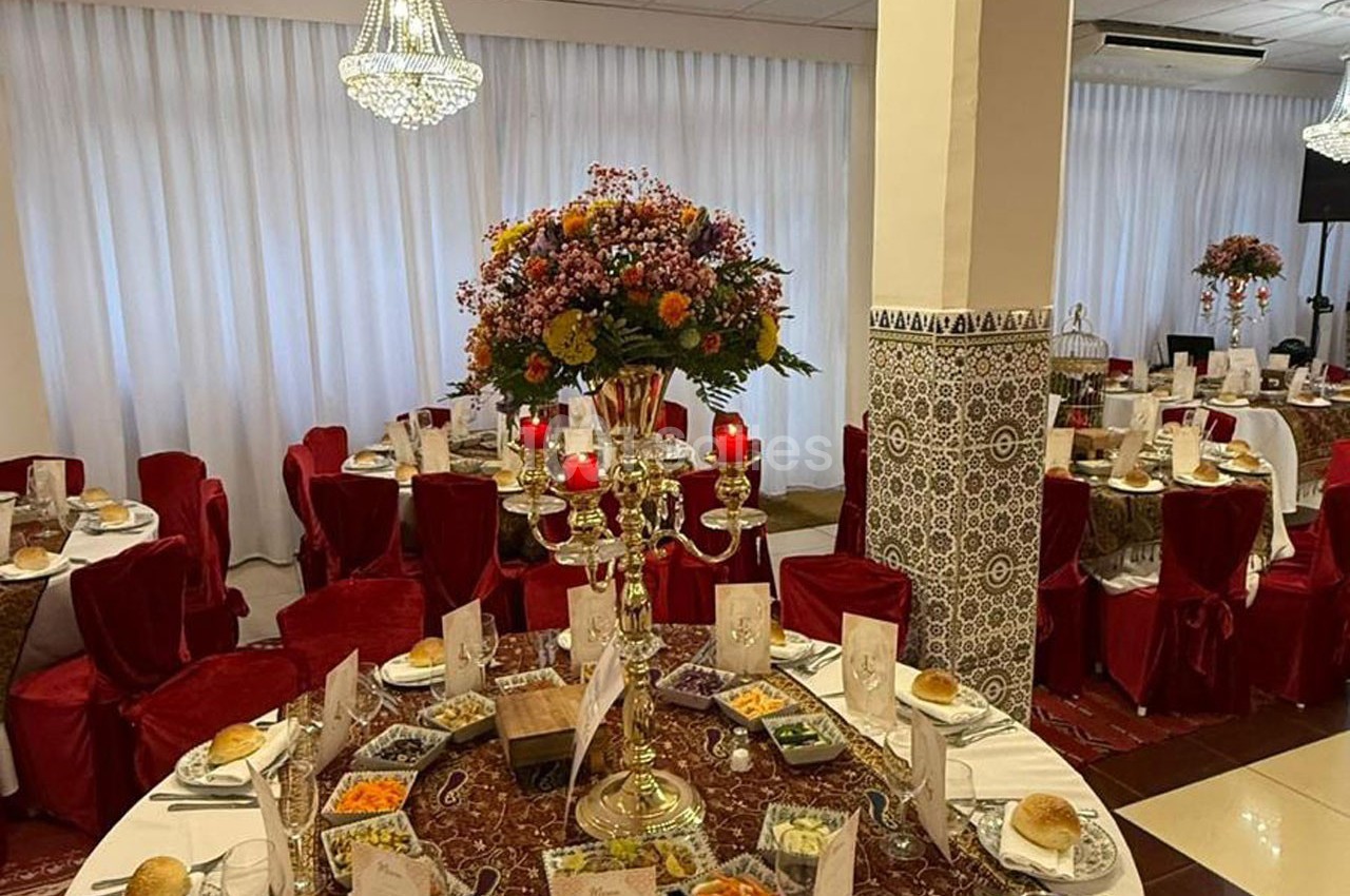 Salle de réception décorée avec des tables dressées, nappes rouges, chandeliers dorés et un grand bouquet floral.