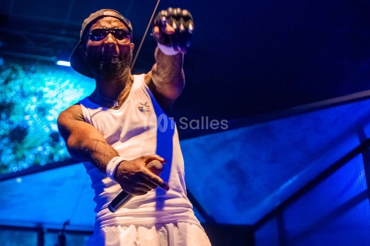 Un chanteur sur scène, tenant un micro, vêtu d'un débardeur blanc et d'une casquette, éclairé par des lumières bleues.