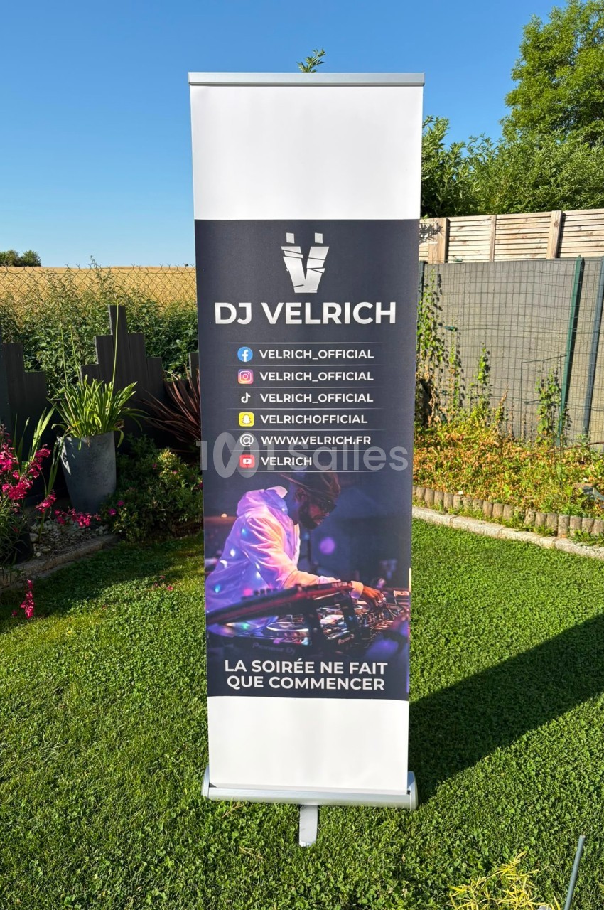 Roll-up publicitaire avec le logo ’DJ Velrich’, des réseaux sociaux et un DJ mixant, installé dans un jardin ensoleillé.