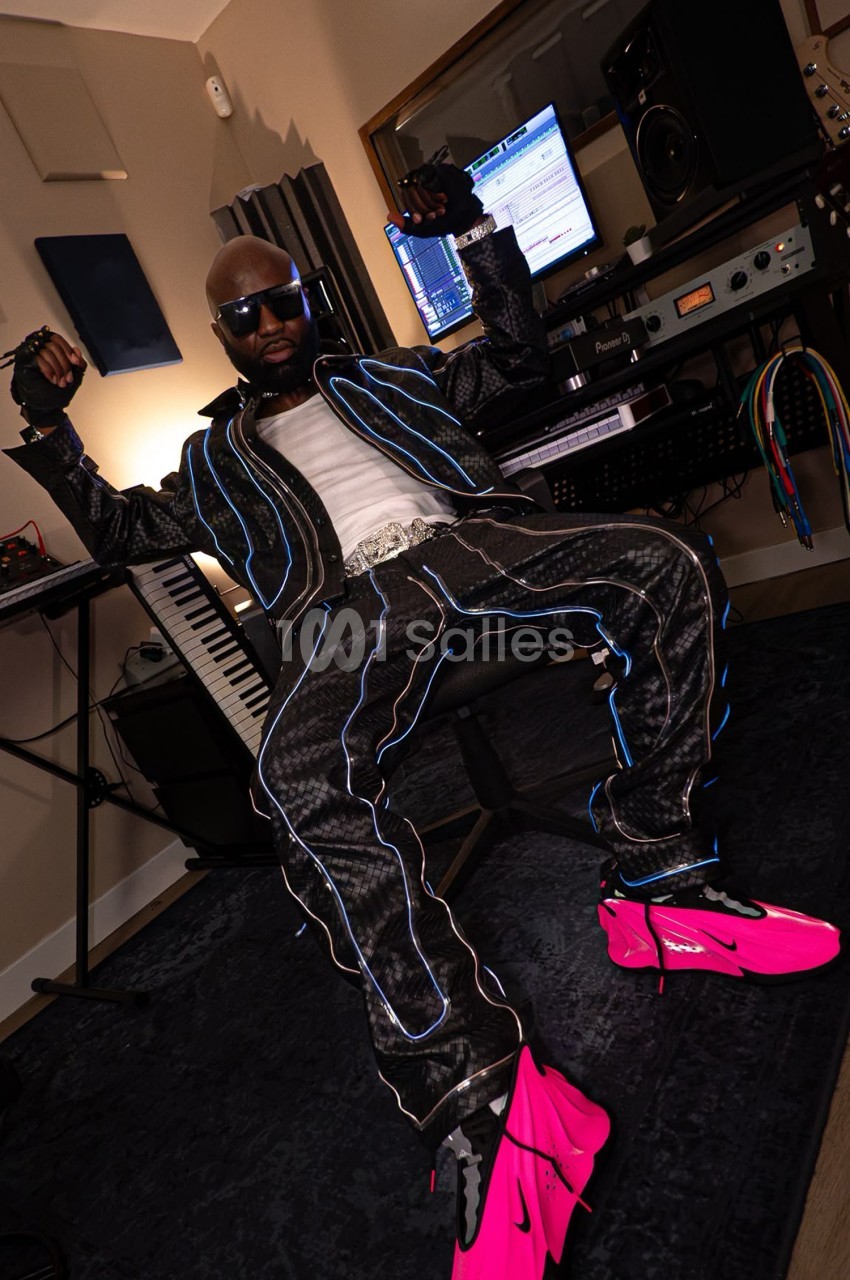 Un homme assis dans un studio d'enregistrement, portant une tenue noire avec des motifs lumineux et des chaussures roses.