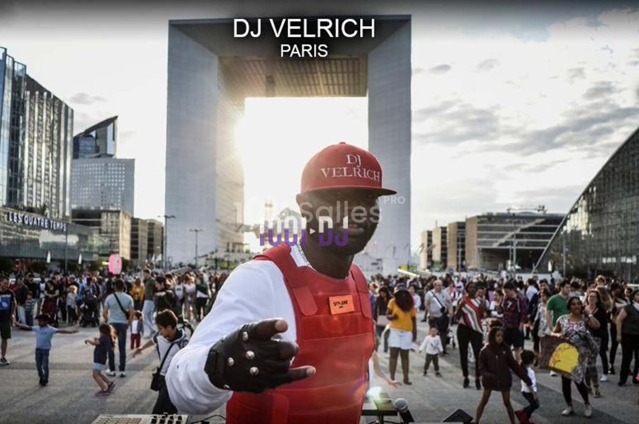Un DJ mixant en plein air devant une foule, avec l'Arche de la Défense en arrière-plan au coucher du soleil.