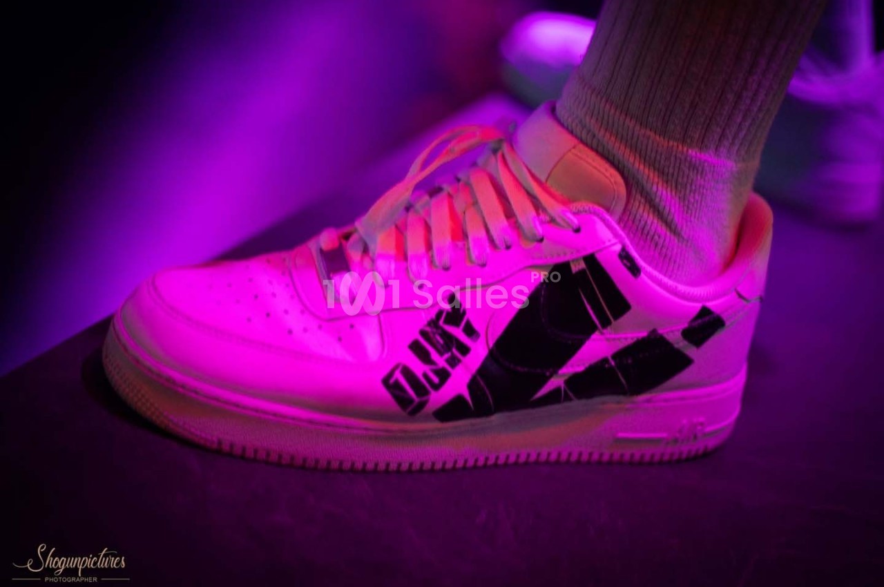 Une chaussure de sport blanche avec des motifs noirs, éclairée par une lumière violette, portée avec une chaussette blanche.