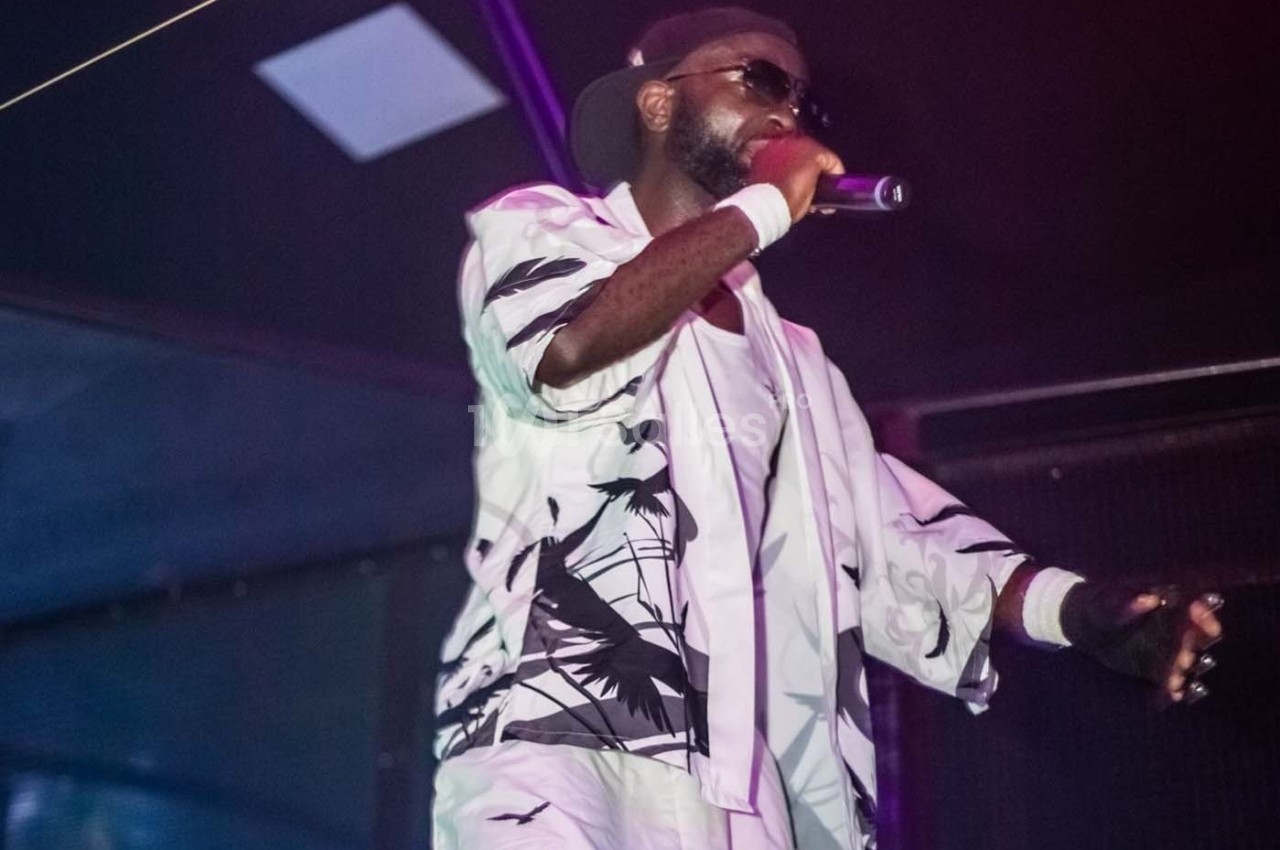 Un chanteur sur scène, vêtu de blanc avec des motifs noirs, tenant un micro et portant des lunettes de soleil.
