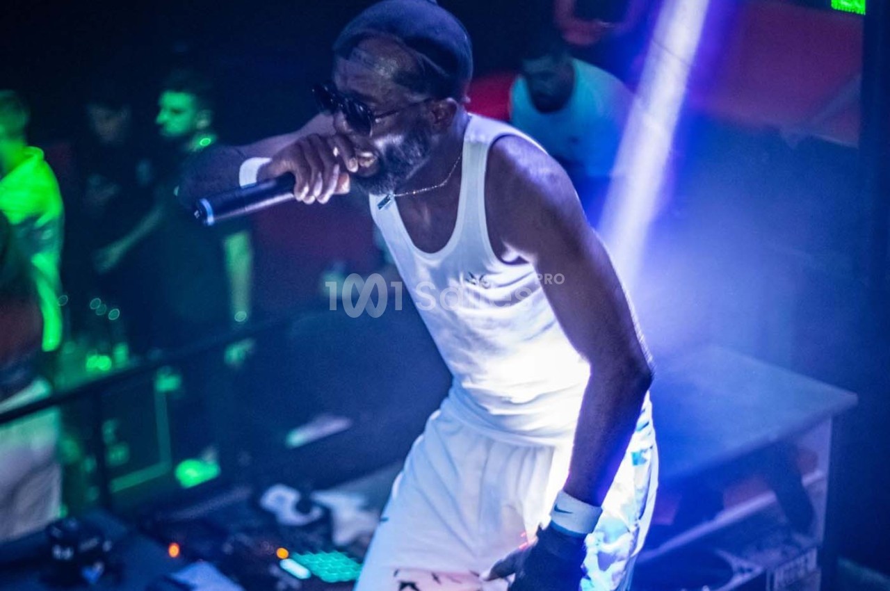 Un chanteur sur scène, vêtu de blanc, tenant un micro, éclairé par des lumières vives dans une ambiance de concert.