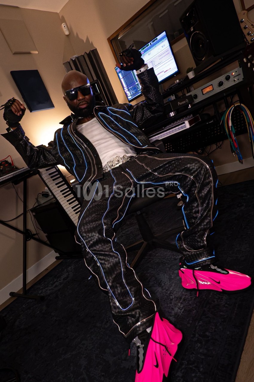 Un homme assis dans un studio d'enregistrement, portant une tenue noire avec des motifs lumineux et des chaussures roses.
