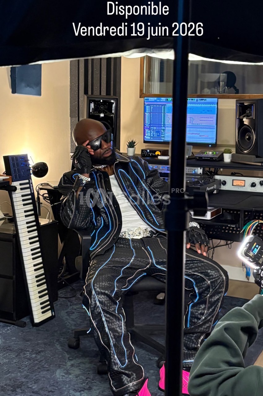 Un artiste en studio d'enregistrement, vêtu de vêtements stylés, assis près d'un clavier et d'équipements audio.