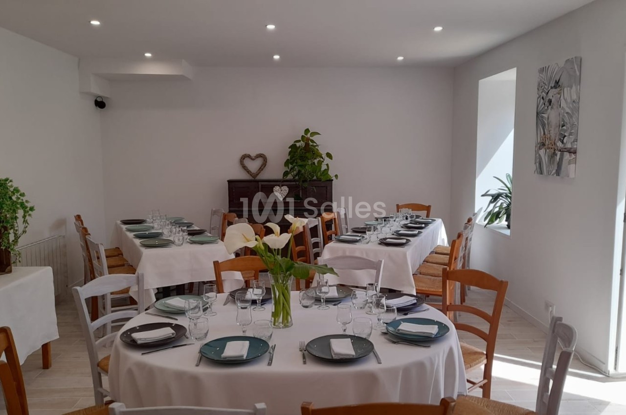 Salle lumineuse avec tables dressées, nappes blanches, chaises en bois, plantes et décoration sobre.