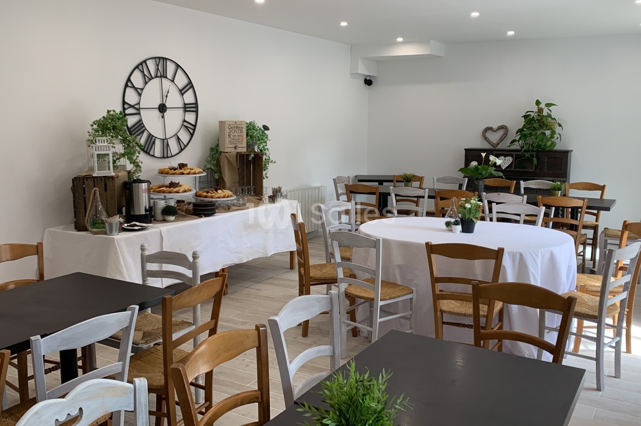 Salle lumineuse aménagée avec des tables et chaises en bois, buffet décoré de plantes et horloge murale.