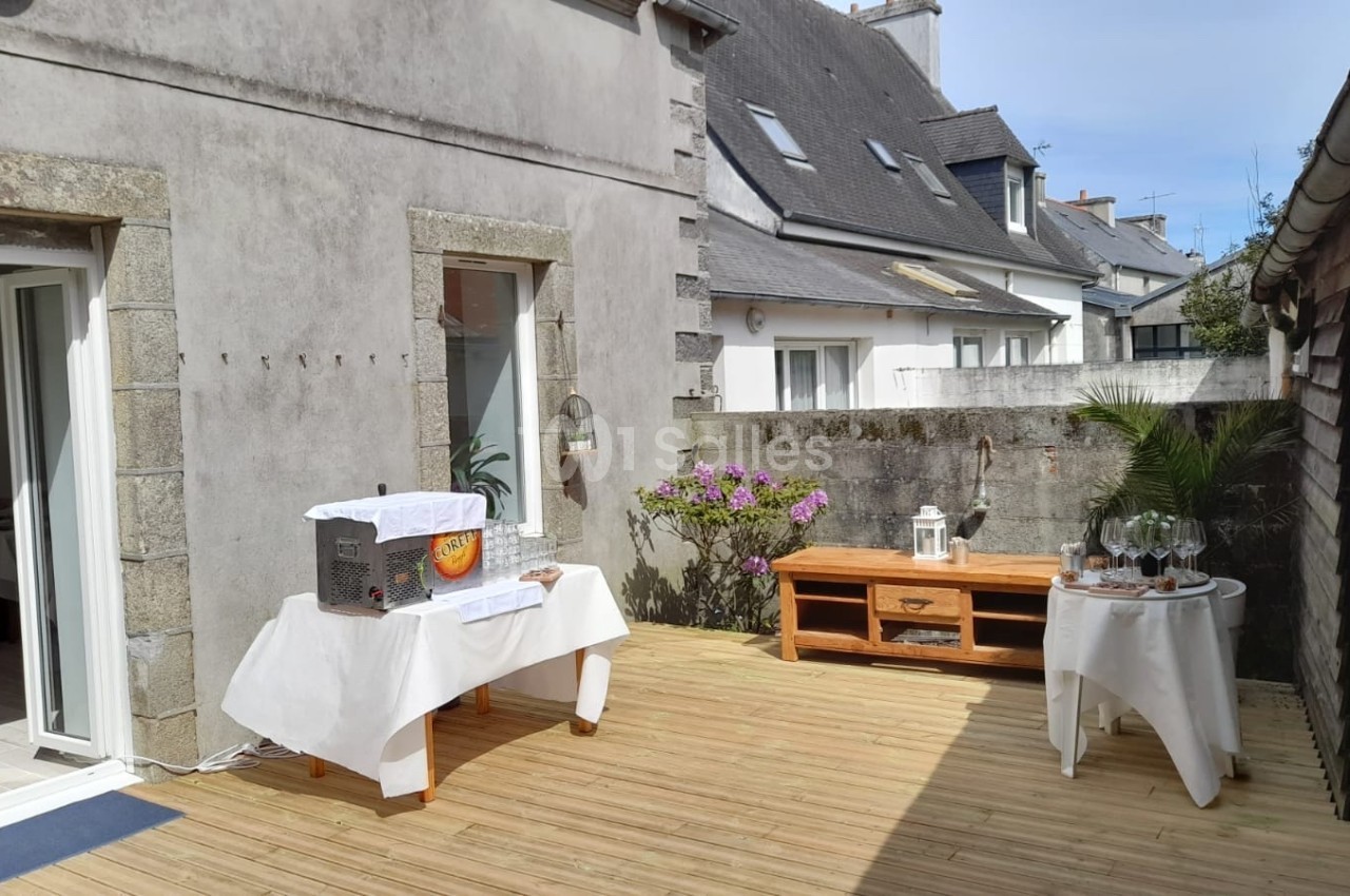Terrasse en bois aménagée avec des tables décorées, entourée de murs en pierre et de maisons voisines.