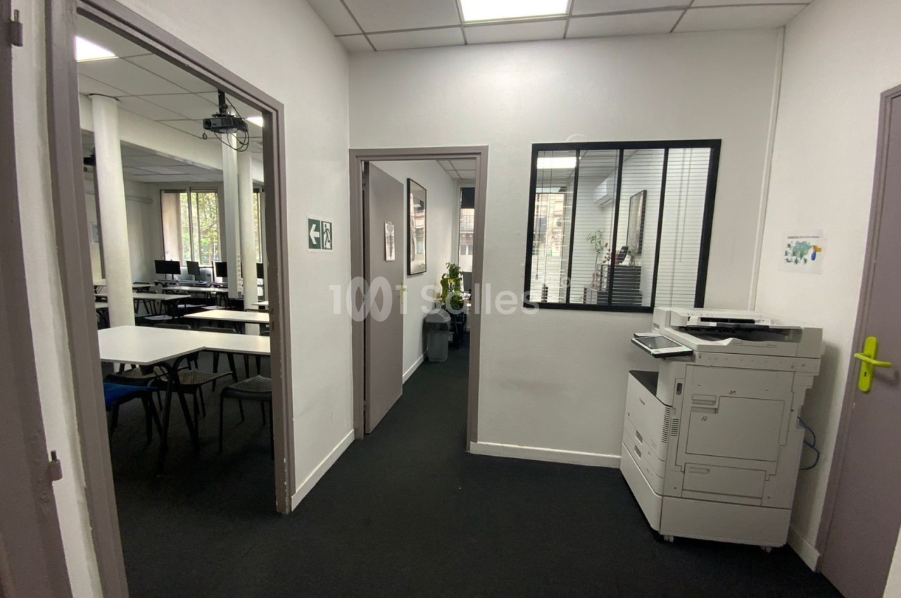 Couloir d'un espace de bureaux avec une imprimante à droite et une salle de réunion visible à gauche.