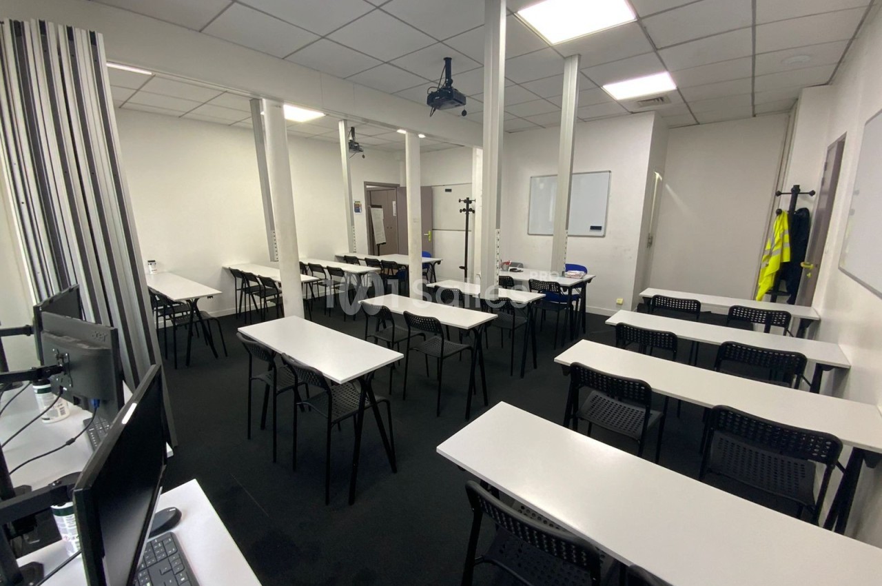 Salle de classe vide avec tables blanches, chaises noires, tableau blanc et projecteur au plafond.