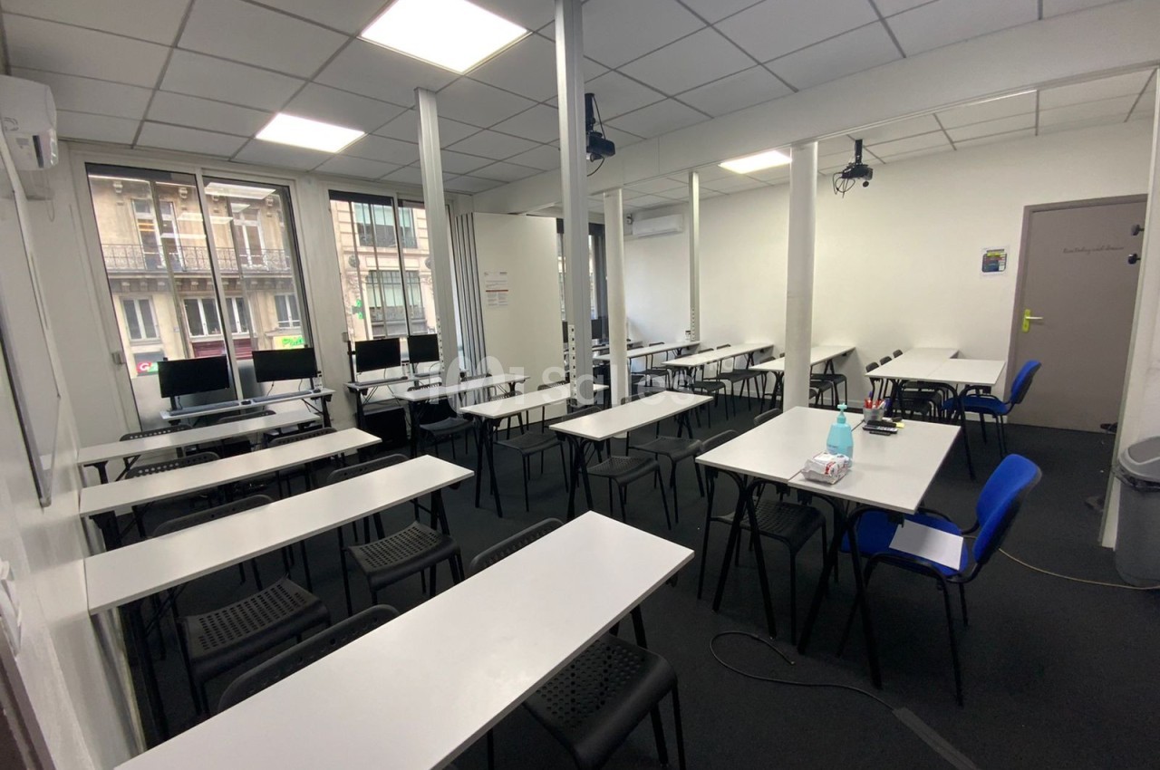 Salle de formation lumineuse avec des tables blanches, des chaises noires et des ordinateurs près des fenêtres.