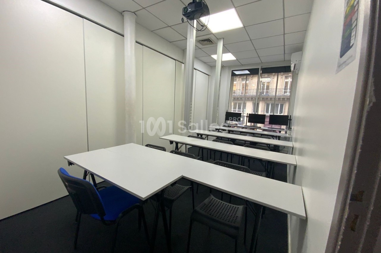 Salle de formation équipée de tables, chaises, projecteur au plafond et ordinateurs près d'une fenêtre.