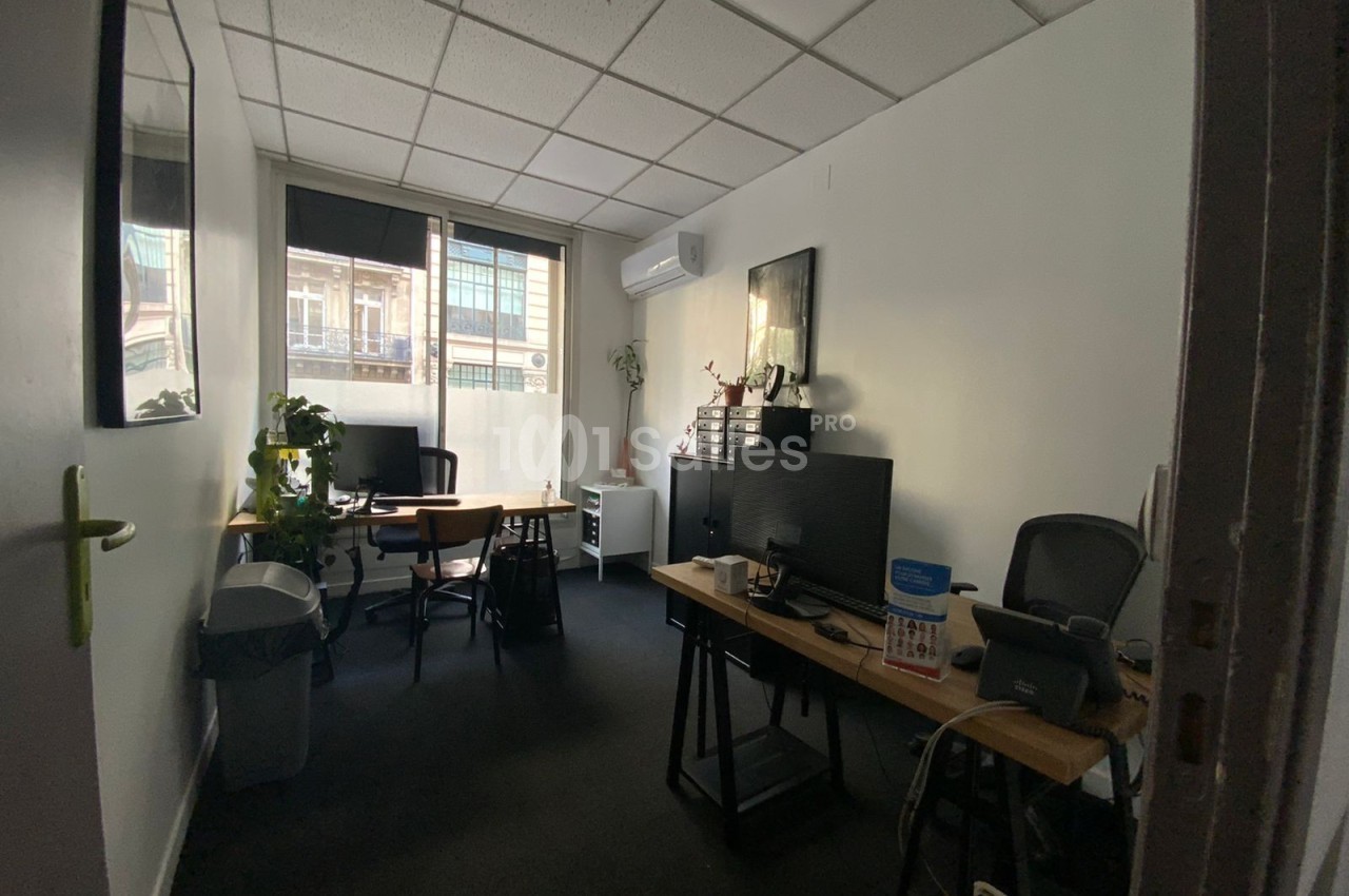 Bureau lumineux avec deux postes de travail, des plantes, des étagères et une fenêtre donnant sur des immeubles.