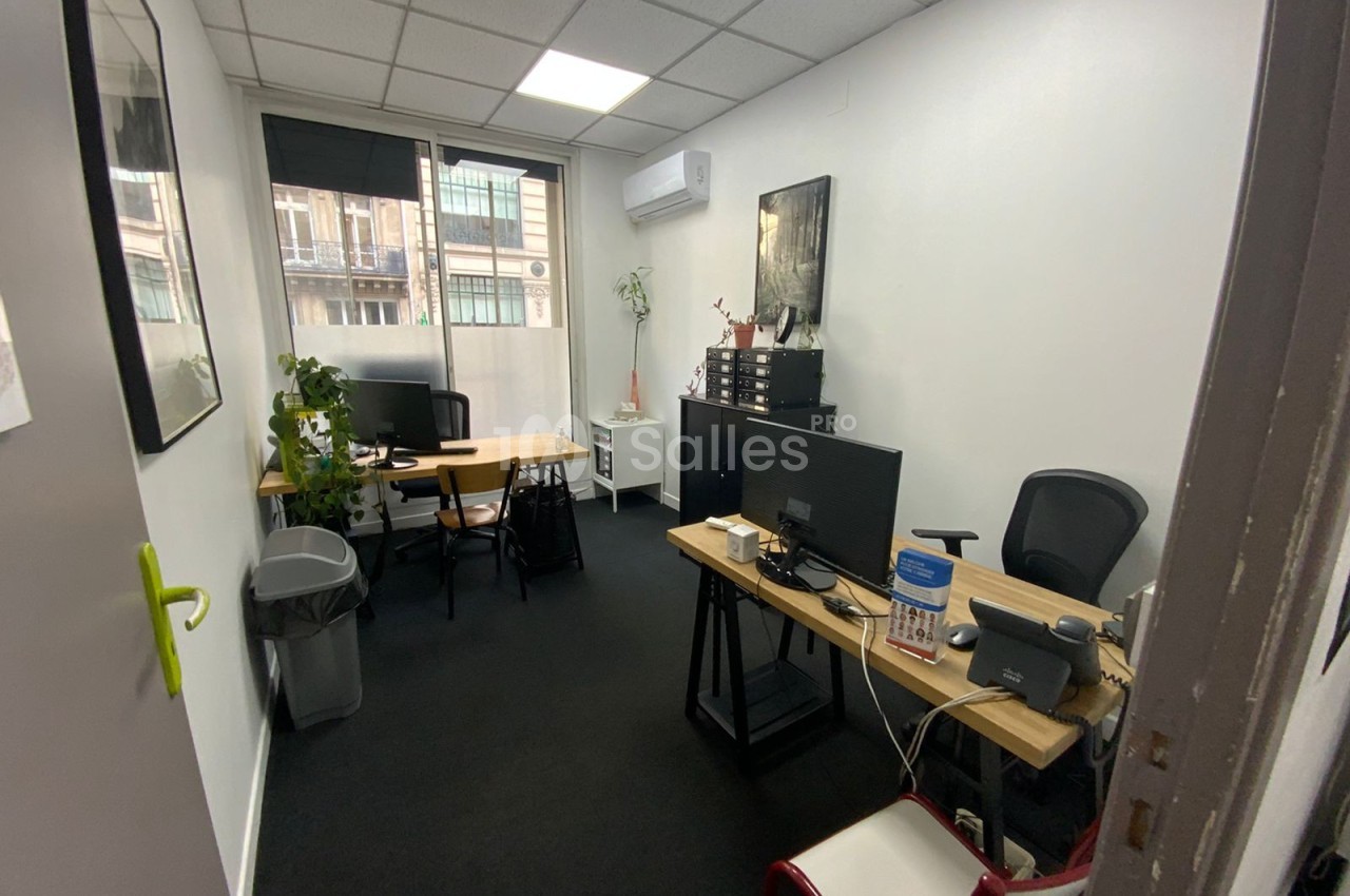 Bureau lumineux avec deux postes de travail, plantes, mobilier moderne et fenêtre donnant sur des bâtiments urbains.