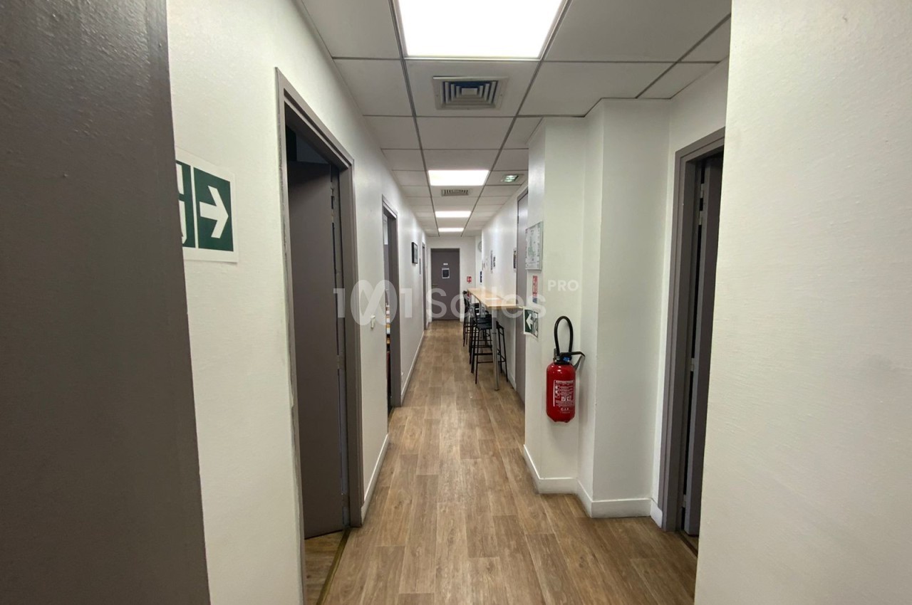 Couloir lumineux avec sol en bois, portes fermées, extincteur mural et signalisation de sortie sur la gauche.